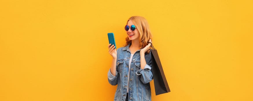 Beautiful happy young woman looking at mobile phone with black shopping bag on yellow studio background