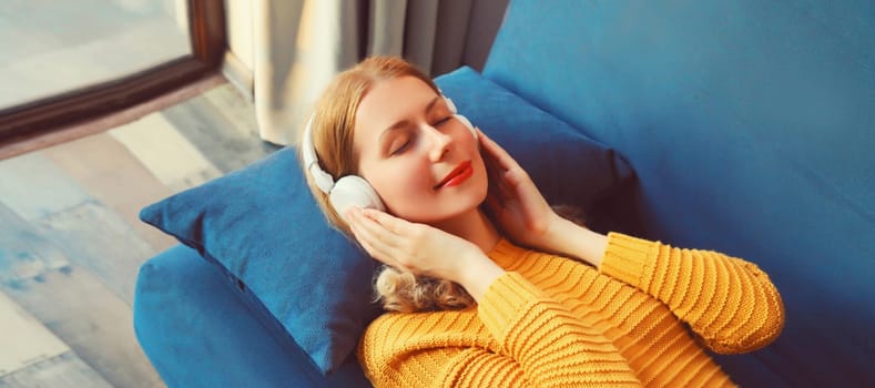 Happy relaxed young woman listening to music with wireless headphones with closed eyes lies on the couch at home