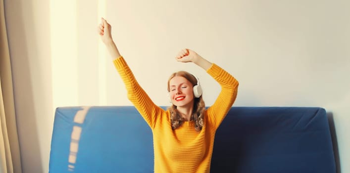Happy cheerful young woman listening to music with headphones having fun sitting on sofa at home
