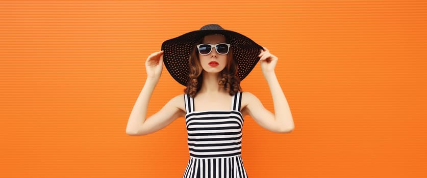 Portrait of beautiful young woman posing wearing black summer straw hat and dress on orange background