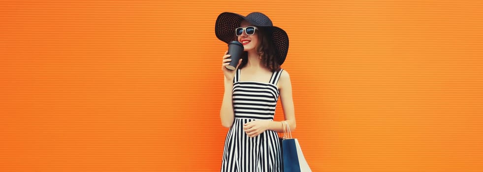 Stylish beautiful happy young woman model with shopping bags in summer black straw hat, striped dress on orange background