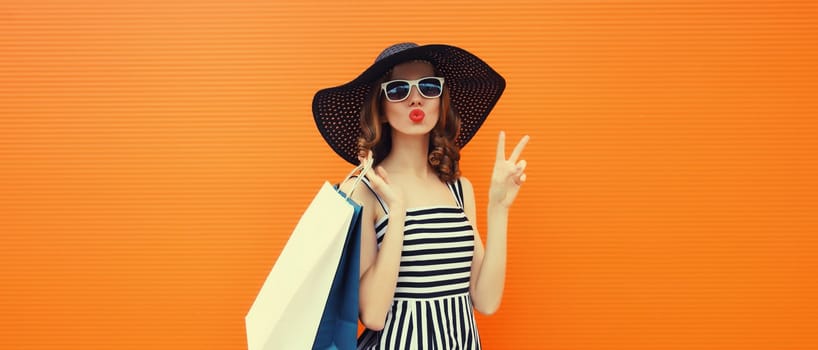 Stylish beautiful happy young woman model with shopping bags in summer black straw hat, striped dress on orange background