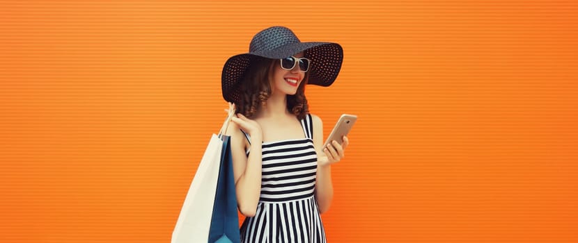 Beautiful happy young woman looking at phone with shopping bag in summer black straw hat, striped dress on orange background