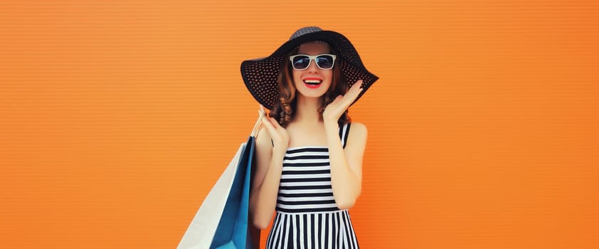 Stylish beautiful happy young woman model with shopping bags in summer black straw hat, striped dress on orange background