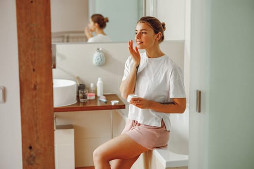 Beautiful young woman taking care of skin by applying moisturizer cream in bathroom