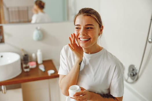 Happy woman applying hydrating moisturizer on her face sitting in bathroom. Home beauty routine