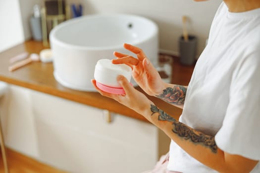 Close up of woman opening jar with face cream while standing in bathroom. Home beauty routine
