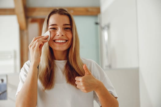 Young woman removing makeup with cotton pad standing in bathroom. Home beauty routine
