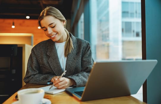 Pretty businesswoman making notes and browsing laptop sitting in modern coworking