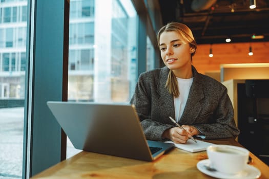 Young female student is making notes and browsing laptop sitting in cafe near window