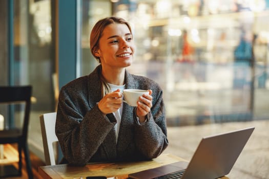 Smiling female entrepreneur is drinking coffee in coworking while working on laptop near window