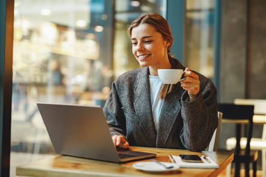 Stylish female freelancer is drinking coffee in cafe while working on laptop near window