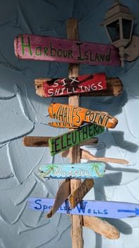 Colorful rustic signpost against a blue wall, featuring Bahamian destinations, under a white lamp in a sunny tropical setting.