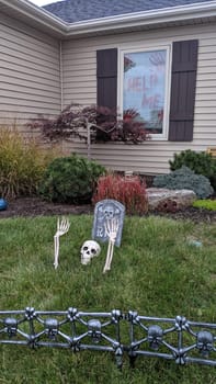 Spooky suburban home decorated for Halloween, featuring a mock graveyard, skeletal remains, and a chilling 'HELP ME' message in the window.