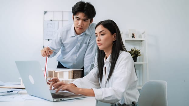 Professional male asian architect using ruler to measure house model length while young beautiful caucasian colleague using laptop to analyzed data on meeting table with house model. Immaculate.