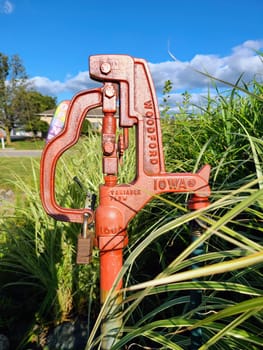 Vintage Woodford water pump in vibrant Iowa setting, secured with padlock amidst lush greenery under clear blue sky