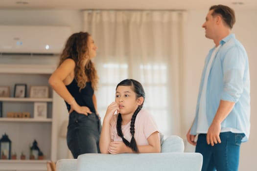 Annoyed and unhappy young girl sitting on sofa trapped in middle of tension by her parent argument in living room. Unhealthy domestic lifestyle and traumatic childhood develop to depression.Synchronos