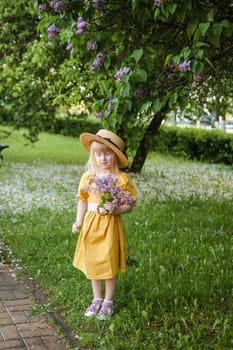 A little girl in a yellow dress and straw hat wearing a bouquet of lilacs. A walk in a spring park, blossoming lilacs