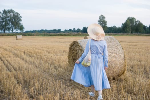 A red-haired woman in a hat and a blue dress walks in a field with haystacks