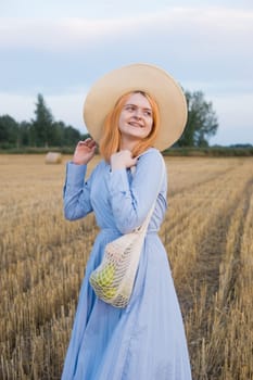 A red-haired woman in a hat and a blue dress walks in a field with haystacks