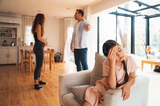 Annoyed and unhappy young girl sitting on sofa trapped in middle of tension by her parent argument in living room. Unhealthy domestic lifestyle and traumatic childhood develop to depression.Synchronos