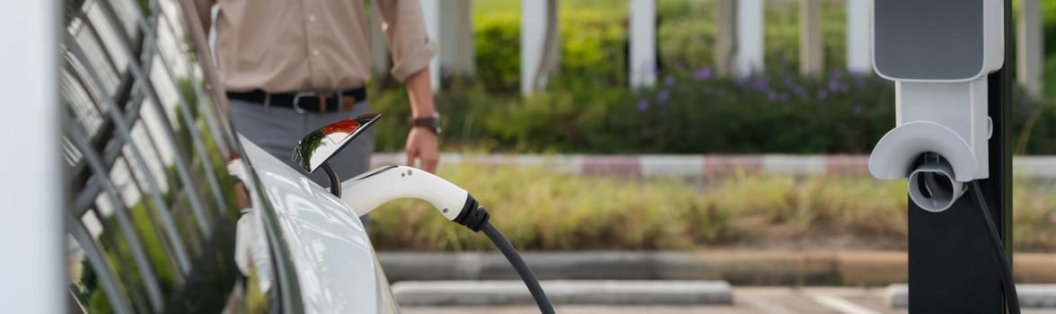 Young man recharge electric car's battery from charging station in city commercial parking lot. Rechargeable EV car for sustainable environmental friendly urban travel. Panorama Expedient