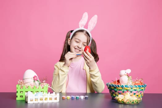 Little enthusiastic child drawing colorful designs and painting eggs with watercolors to prepare for the Easter festivities, pink backdrop. Young small girl relaxing as she creates art at table.