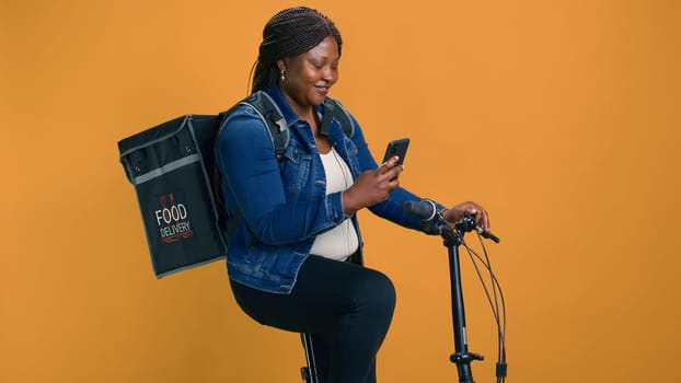 African american female multitasking on her bike, delivering food with cellphone in hand. Youthful black woman checking delivery app on mobile device for efficient transportation of orders.