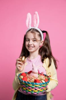 Small positive kid with bunny ears shows Easter toys and items, presenting exciting april festivities. Young cheerful girl with adorable pigtails showing a rabbit in a basket and colorful eggs.