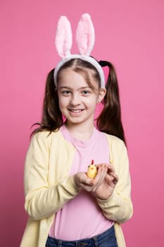 Little smiling girl holding an adorable stuffed chick in front of camera, cheerful young kid with fluffy golden toy over pink background. Small child with cute bunny ears for festivity.