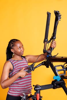 Determined african american lady adjusting and securing bicycle headset and handlebar in front of yellow background. Disassembled bike being repaired by sports-loving female for leisure cycling.