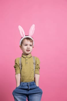 Young adorable boy smiling and standing over pink backdrop, wearing bunny ears to celebrate easter festivity. Cheerful small kid being happy in front of camera, waiting to receive presents.