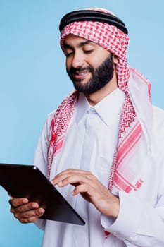 Smiling muslim person dressed in traditional clothes using digital tablet while engaging with social media messages. Cheerful arab person in islamic thobe and headdress using portable device in studio
