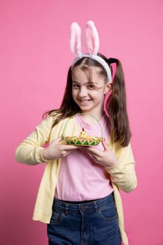 Lovely little kid with rabbit ears poses with adorable springtime toys while carrying an Easter basket full of eggs and a chick. Joyful and enthusiastic youngster enjoying the holidays in studio.