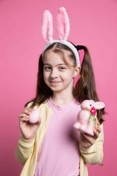 Lovely positive kid holding fluffy cute ornaments in front of camera, presenting her easter toys and decorations. Joyful young kid wearing bunny ears and smiling over pink background.