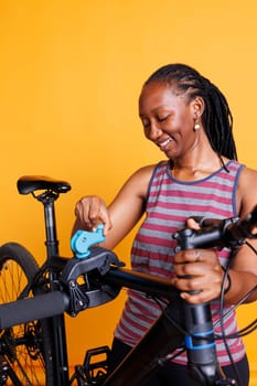 African american female cyclist inspects her broken bicycle frame on repair stand against isolated yellow backdrop. Black woman preparing to make adjustments and repairs with specialized equipment.
