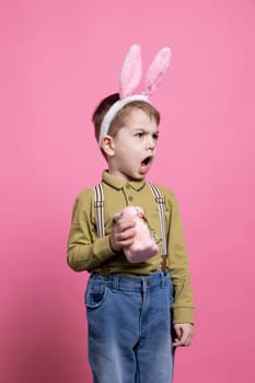 Adorable little kid being upset and unhappy in front of camera, holding a stuffed handmade rabbit toy against pink background. Young small boy wearing bunny ears and feeling sad.