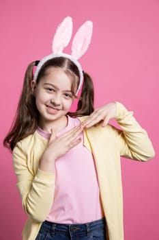 Cute young girl dancing and fooling around on camera, wearing bunny ears and pigtails over pink background. Carefree schoolgirl feeling joyful and excited about easter celebration in studio.