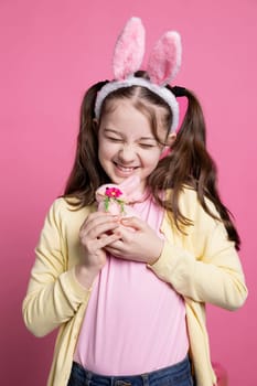 Smiling young child embracing a stuffed rabbit toy in studio, wearing bunny ears and holding easter decoration in front of the camera. Small girl with pigtails being proud of fluffy toy.