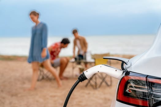 Alternative family vacation trip traveling by the beach with electric car recharging battery from EV charging station with blurred family enjoying the seascape background. Perpetual