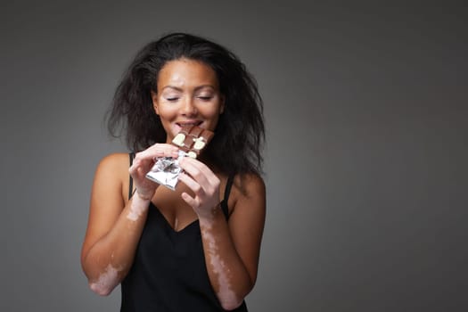 Smiling African American woman with Vitiligo disease eating chocolate. Beautiful young lady with dark hair having sweet food against gray background. Black lady with skin problem.