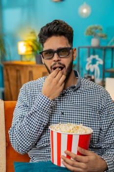 Indian Hindu man taking break from work wearing 3D glasses eating popcorn watching movie film content on TV. Smiling Hispanic guy sits at home couch sofa having snacks during weekends. Vertical
