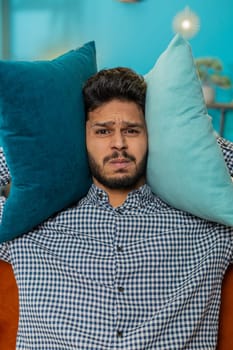 Repair work at neighbors. Irritated Indian Hindu man cover ears with pillows annoyed by noisy loud music neighbors suffer from headache wishes silence. Thin walls at home flat without sound insulation