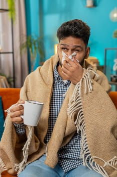Indian ill man suffering from cold or allergy lying on sofa in living room at home. Sick Hispanic guy with towel on head blows sneezes wipes snot into napkin. Coronavirus quarantine pandemic. Vertical