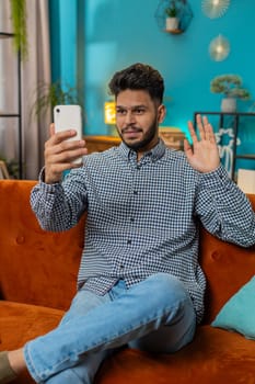 Happy Indian Arabian man blogger taking selfie on smartphone, communicating video call online with subscribers recording stories for social media. Hispanic guy sitting at home orange couch. Vertical