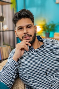 Portrait of happy Indian young man at home couch smiling friendly, glad expression looking away dreaming resting, relaxation feel satisfied good news, celebrate win. Hispanic adult guy in living room