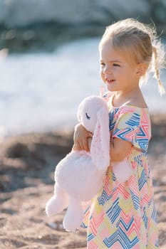 Little smiling girl with her hair fluttering in the wind stands on the beach with a plush bunny and looks at the sea. High quality photo