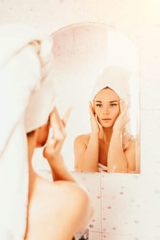 Young beautiful woman using face cream moisturizing lotion after bath. Pretty attractive girl in a towel on her head stands in front of a mirror in a home bathroom. Daily hygiene and skin care.