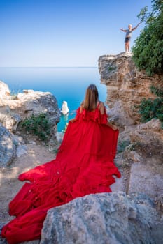 Red dress sea woman. Happy woman with flowing hair in a long flowing red dress stands on a rock near the sea. Travel concept, photo session at sea
