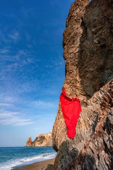 woman sea red dress. Woman with long hair on a sunny seashore in a red flowing dress, back view, silk fabric waving in the wind. Against the backdrop of the blue sky and mountains on the seashore